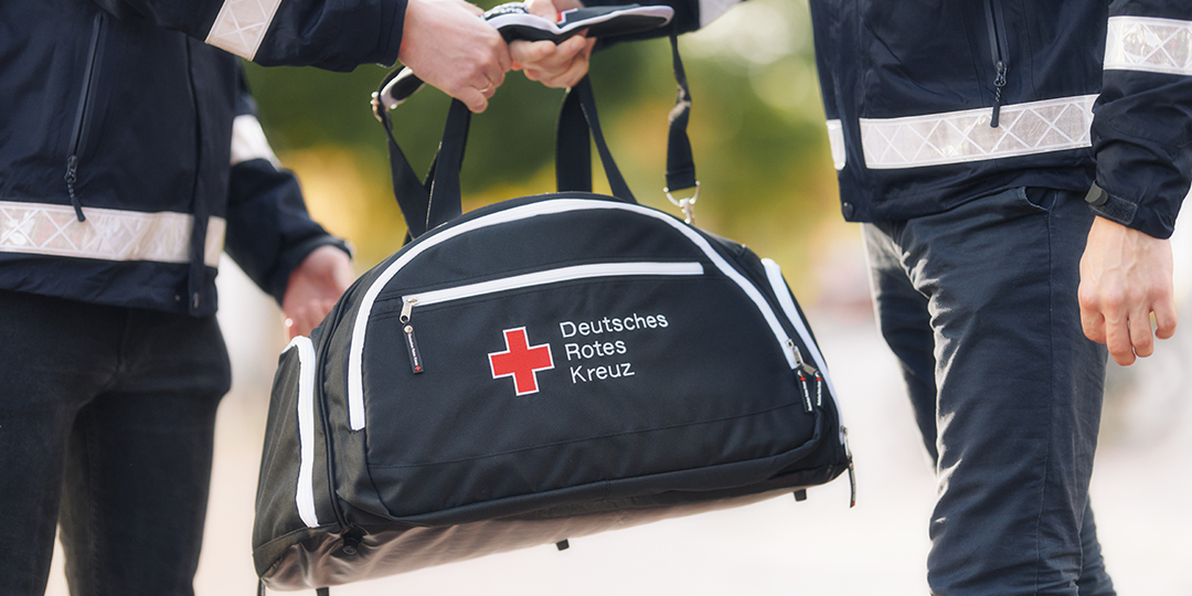 Zwei Personen halten gemeinsam eine schwarze Sporttasche im Retrolook mit gesticktem DRK-Kompaktlogo